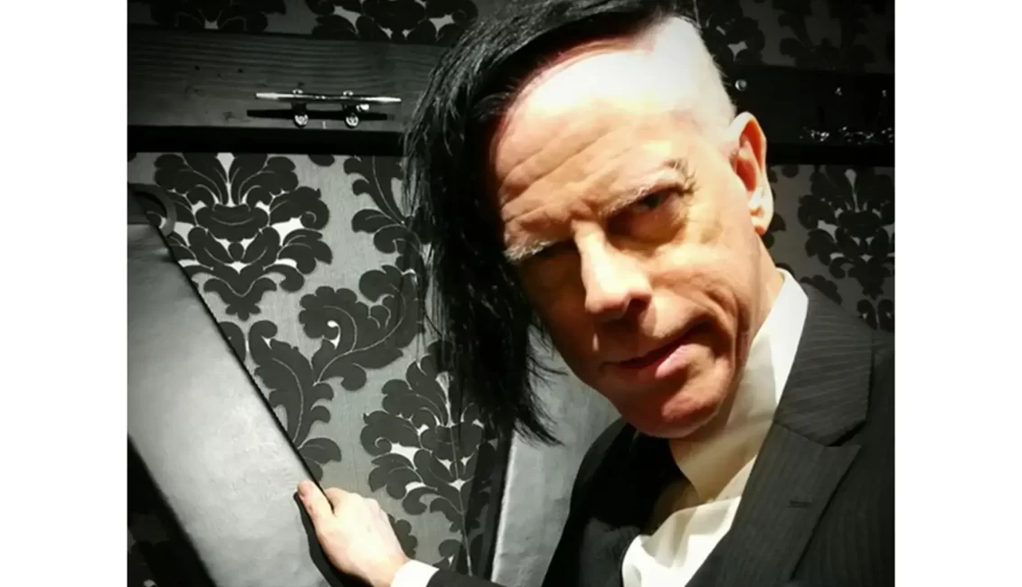 An image of Danarama from Kink.com facing forward with his hand on a St. Andrews Cross and facing towards the camera