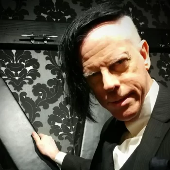 An image of Danarama from Kink.com facing forward with his hand on a St. Andrews Cross and facing towards the camera