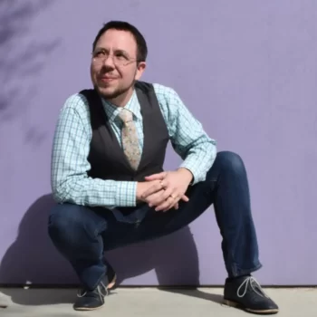 An image of Lee Harrington from passion and soul wearing a straw hat and holding a wooden staff with a peacock feather and beads hanging from it. He is looking at the camera and smirking
