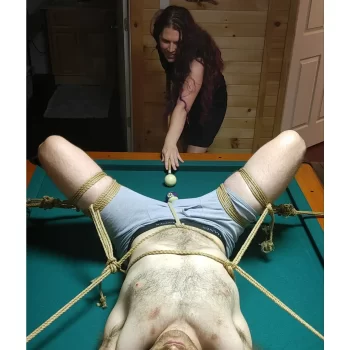 An image of IPCookieMonster SlutPhD smiling as she plays pool and aims her cue to hit a billiard ball aimed at her male submissive's crotch. He is lying on the pool table in only underwear and he is tied to the table spread-eagled