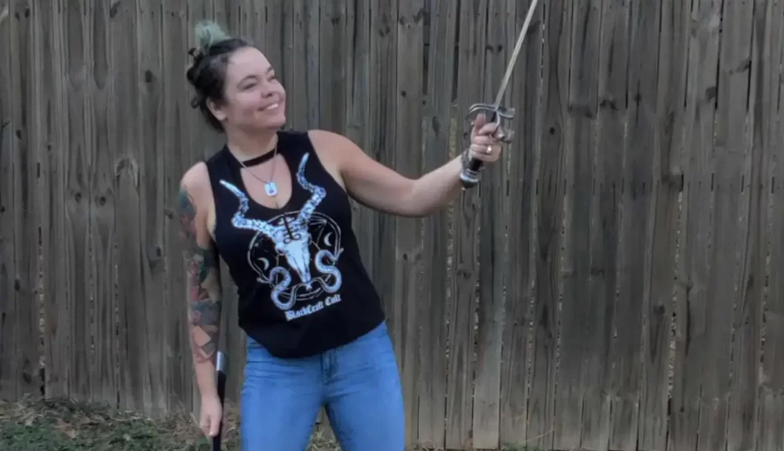 An image of Farrah Bonnot standing outside wielding a sword and smiling. She is wearing a black tank top and jeans with black boots.