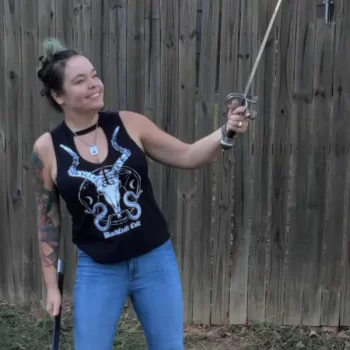 An image of Farrah Bonnot standing outside wielding a sword and smiling. She is wearing a black tank top and jeans with black boots.