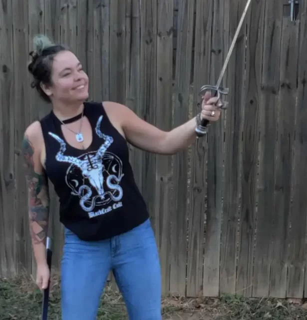 An image of Farrah Bonnot standing outside wielding a sword and smiling. She is wearing a black tank top and jeans with black boots.