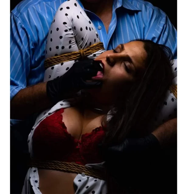 An image of Nkay Nadia. She is tied in rope and her shirt is open to reveal red lingerie. Niho's hands are in her mouth and pulling her back towards him.